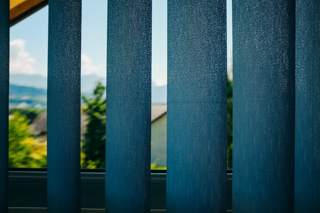 Through blue blinds view on green trees growing in yard of home located in suburb